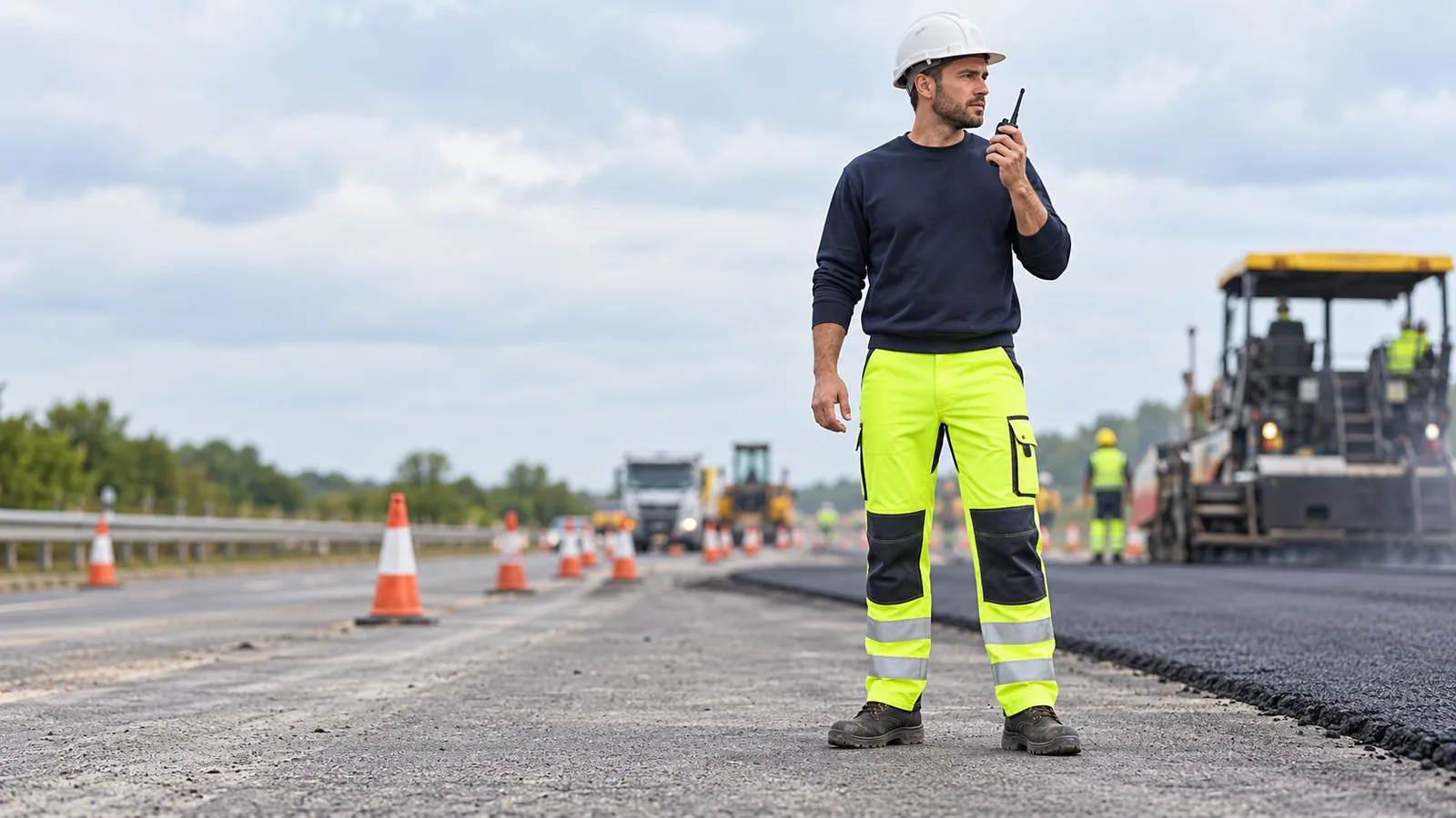 Full-length view of a worker wearing fluorescent yellow-green hi vis work trousers with leg-level reflective bands walking through a daytime road work zone